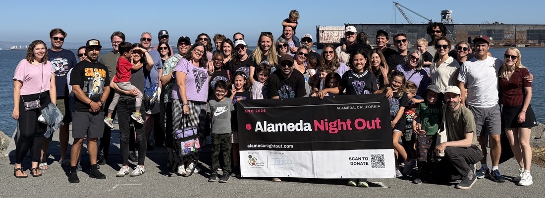 Around forty Alameda neighbours behind the Alameda Night Out banner on the shoreline at Alameda Point.