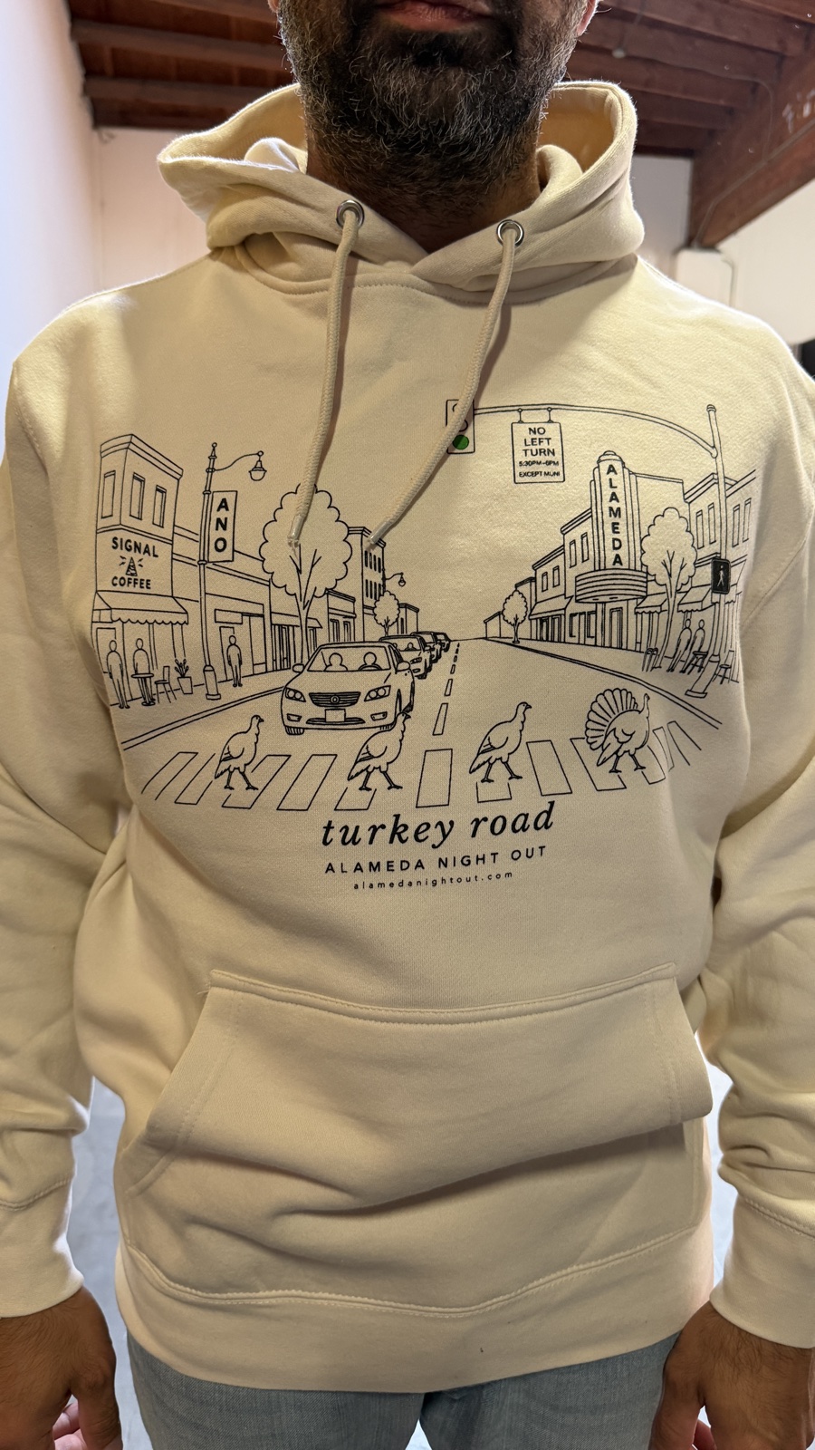 The Turkey Road Hoodie in cream: dark line art of the Alameda theater and turkeys crossing the street